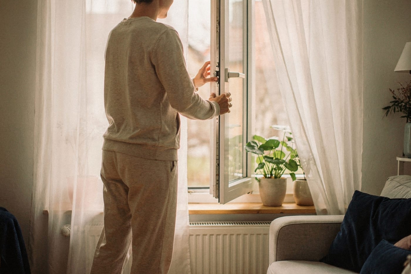 Person öffnet in einem hellen Wohnzimmer das Fenster zum Lüften, Zimmerpflanzen auf der Fensterbank – symbolische Darstellung